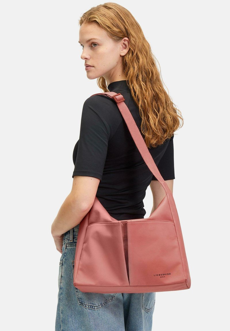 Różowa, luźna torba na ramię wykonana z gładkiego materiału, z dwoma przednimi kieszeniami i długim, regulowanym paskiem do wszechstronnego noszenia.