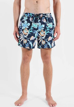 Homme portant un short de bain bleu marine avec des motifs floraux tropicaux bleu clair et orange, debout pieds nus devant un fond clair uni.