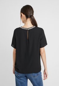 Top corto de manga corta negro con un escote decorativo de cuentas y una abertura en la parte posterior en forma de llave. La tela parece suave y ligera.