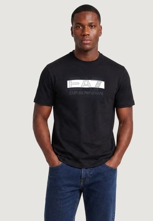 Uomo con capelli corti che indossa una t-shirt nera con il logo Emporio Armani EA7 e jeans blu, in piedi con le mani in tasca, sfondo neutro.