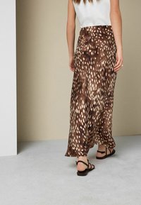 Femme qui s'éloigne portant un top blanc sans manches, une longue jupe marron à motif animalier et des sandales plates noires sur un fond uni.