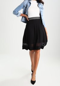 Mujer con una blusa blanca, chaqueta de mezclilla, falda negra con dobladillo de encaje y tacones negros, posando con una mano en la cadera.