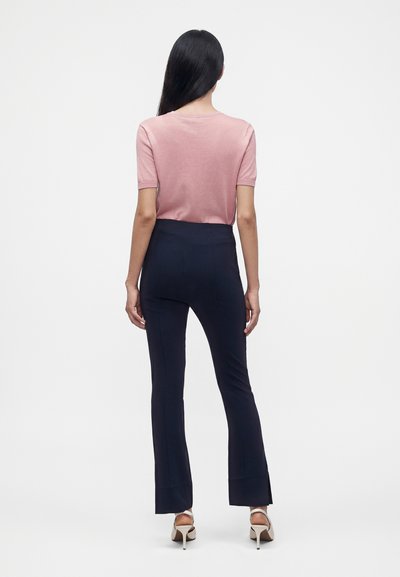 Femme aux longs cheveux noirs, portant un haut rose à manches courtes, un pantalon taille haute bleu marine et des sandales blanches à talons hauts, debout de dos.
