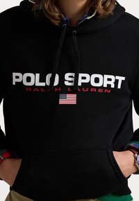 Schwarzer Hoodie mit "POLO SPORT" und "RALPH LAUREN" in Weiß und Rot gedruckt. Mit einem Patch der amerikanischen Flagge und Kapuze mit Kordelzug.