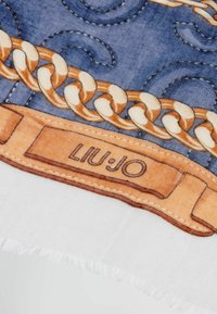 Foulard de seda con un fondo azul, un patrón de cadenas doradas y una etiqueta naranja con "LIU JO" impresa. Los bordes están deshilachados.