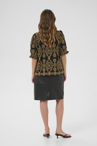 Svart blus med gyllene mönstrad broderi, puffärmar och vågig nederkant, tillsammans med en figursydd svart läderskjort. Platta svarta sandaler.