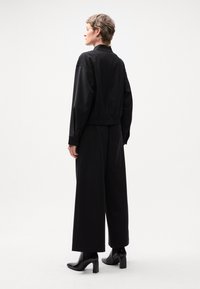 Veste noire à col montant et ceinture élastique, associée à un pantalon noir à jambes larges. Le modèle porte des bottes noires à talons.