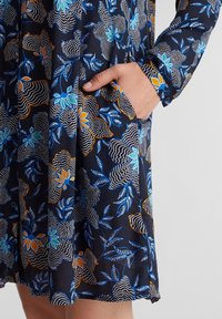 Abito a maniche lunghe con sfondo blu navy e motivo floreale nei toni del blu, arancio e bianco. Include una tasca laterale e dettagli con bottoni.