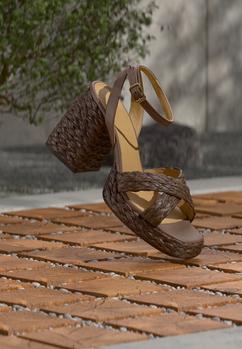 Sandale à talon haut tressée marron avec bride à la cheville, flottant au-dessus de carreaux en bois à motifs en plein air près de la végétation et d'un mur en béton.