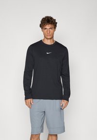 Sort langærmet skjorte lavet af bomuld, med et hvidt Nike-logo på brystet. Kombineret med grå sportsbukser.