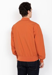 Orange Bomberjacke mit einem gerippten Saum und Bündchen, die ein einfaches Design und eine glatte Textur aufweist. Trägt man über dunklen Denim-Jeans.