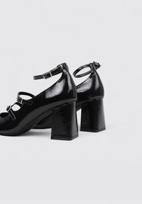 Zapatos de tacón bloque de charol negro que cuentan con correas en el tobillo, lados abiertos con un diseño recortado y un tacón bloque texturizado para mayor estabilidad.