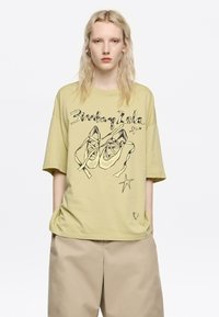 Camiseta de gran tamaño en color amarillo claro, con un dibujo en línea negro de zapatos y estrellas. Fabricada en suave algodón, con un pequeño detalle en forma de corazón en el dobladillo.