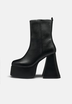 Stivaletto alla caviglia in pelle nera con suola piattaforma spessa e tacco alto largo a zampa d'elefante, texture liscia e piccolo logo vicino al bordo superiore.