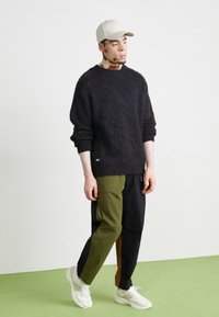 Pullover nero in maglia morbida con maniche larghe, abbinato a pantaloni color block in verde, nero e marrone. Il modello indossa un berretto chiaro e sneakers bianche.
