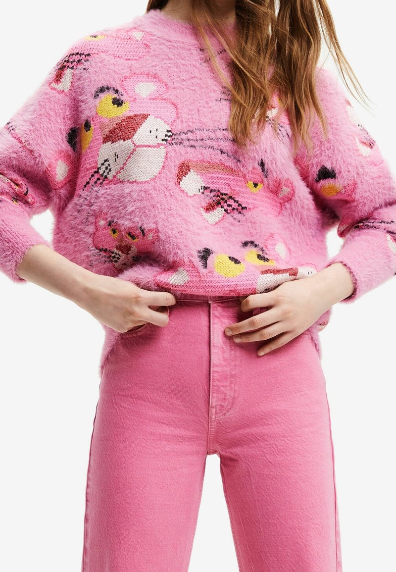 Femme ajustant la taille d'un pantalon rose taille haute, portant un pull rose pelucheux avec des visages de panthère dessinés en jaune et noir.