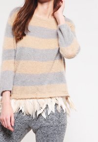 Gestreifter Pullover in Beige und Grau, aus weichem Material, mit einem strukturierten Federbesatz am Saum. Kombiniert mit grauen Strickhosen.