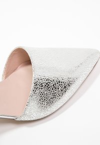 Gros plan d'une chaussure plate argentée texturée à bout pointu avec une découpe sur le côté et une doublure intérieure rose clair sur fond blanc.