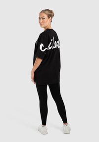 Camiseta negra oversized con la palabra "Chill" en texto cursivo blanco en la parte posterior, combinada con leggings negros y zapatillas blancas. Textura de tela suave.