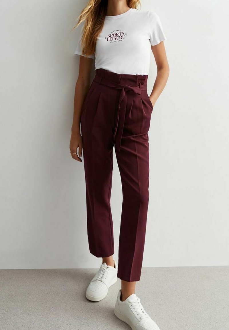 Femme portant un t-shirt blanc "Sports & Leisure", un pantalon bordeaux taille haute avec ceinture et des baskets blanches, debout contre un mur uni.