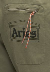 Κοντινή λήψη τσέπης από ελαιώδες πράσινο ύφασμα με μαύρο λογότυπο «Aries», φερμουάρ και πορτοκαλογκρί κορδόνι προσαρτημένο στον μηχανισμό του φερμουάρ.