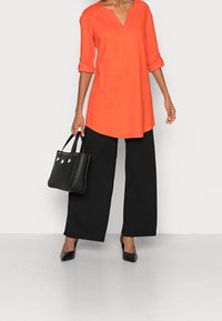 Tunic rosso-arancione con scollatura a V e maniche arrotolate, abbinata a pantaloni neri larghi e tacchi a punta neri, mentre tiene una borsa nera.