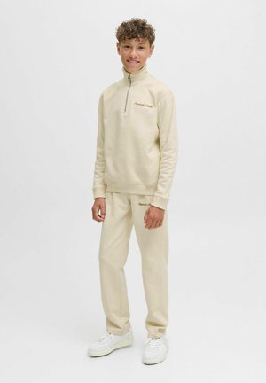 Beige zip-up sweatshirt med hög krage och broderad logotyp, ihopparad med matchande beige joggerbyxor och vita sneakers. Slät textur.