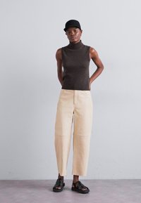 Top marrón sin mangas con cuello alto, pantalones de pierna ancha color crema y sandalias negras. La modelo se encuentra de pie frente a un fondo liso.