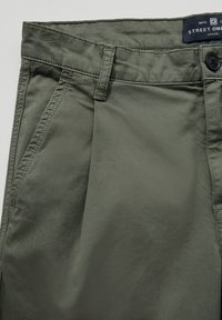 Olivově zelené bavlněné kalhoty s hladkým povrchem. Mají zapínání na knoflík, standardní kapsy a viditelné švy. Viditelný štítek značky.