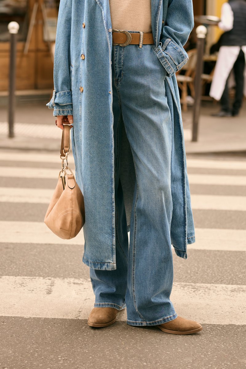 Jeans anchos de mezclilla azul combinados con un abrigo largo azul claro, cinturón marrón y bolso de ante beige, sobre un paso de peatones con pavimento gris.