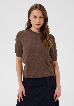 Kvinde med langt brunt hår iført en struktureret brun kortærmet sweater og mørkeblå denimnederdel, stående mod en ensfarvet baggrund.