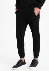 Sorte joggerbukser lavet af blødt stof, med elastiske manchetter, sidelommer og en afslappet pasform, parret med sorte sneakers og en ensfarvet top.