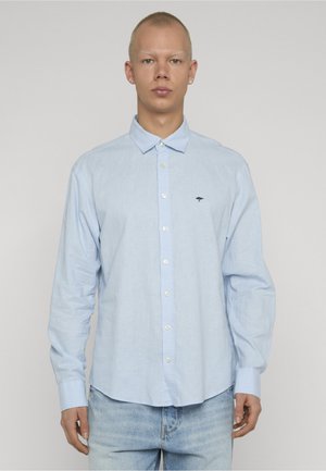 Junger Mann trägt ein hellblaues, langärmliges Hemd mit Knöpfen und kleinem dunklem Logo auf der Brust sowie eine helle Jeans, steht und blickt geradeaus.