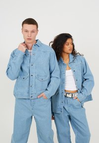 Jachete din denim de culoare albastru deschis cu buzunare frontale și închideri cu capse, purtate cu pantaloni asortați. Una are un broderie decorativă cu logo.