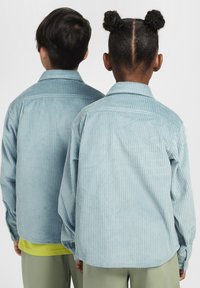 Šviesiai mėlynas kordūro džiazas su vertikaliais įbrėžimais, užsegta apykaklė ir ilgos rankovės, jį dėvi du vaikai iš nugaros.