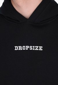 Nahaufnahme eines schwarzen Hoodies mit weißen, gestickten Text "DROPSIZE", der in der Mitte der Brust unterhalb des Halsausschnitts platziert ist.