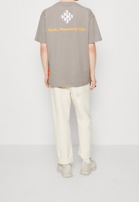 Harmaa ylisuurikokoinen t-paita, jossa on valkoinen ympyrägraafikka ja orange teksti selässä; yhdistettynä kermaiset ylöskäännetyt housut ja beige kengät.