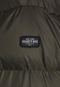 Mørkegrøn, kviltet stof med en sort rektangulær lap, der står "New York SCHOTT NYC Midtown South 1913" broderet på.
