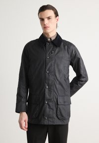 Veste cirée noire avec un col marine, dotée de boutons-pression et de deux poches avant. Sa texture lisse et sa coupe ajustée mettent en valeur son design.