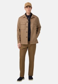 Brun lättviktsjacka med två framfickor, bärs över en marinblå topp, tillsammans med bruna byxor och marinblå slip-on skor. Kapa har logotyp.