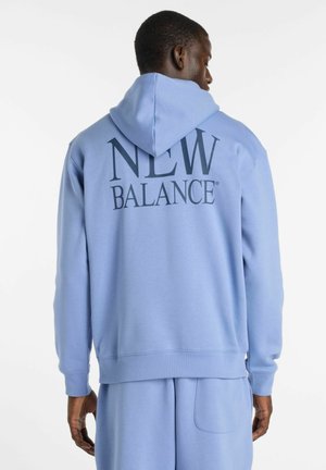 Mann, der von hinten zu sehen ist und eine hellblaue New Balance Kapuzenjacke sowie passende Jogginghosen vor einem einfarbigen weißen Hintergrund trägt.