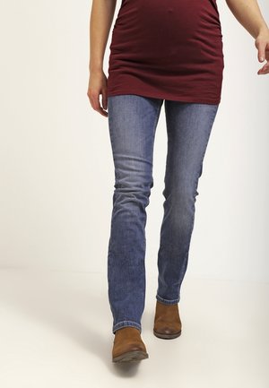 Person, der går, iført blå jeans, brune støvler og en vinrød lang top mod en ensfarvet hvid baggrund.