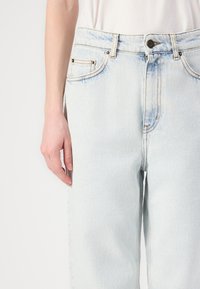 Vaqueros de denim azul claro con un corte relajado, que presentan costuras en contraste, cierre de botón y bolsillos delanteros. Textura lisa, sin patrones visibles.
