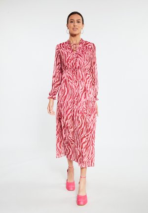 Vrouw in een roze en rode jurk met dierenprint en lange mouwen loopt naar voren met roze plateauhakken tegen een effen witte achtergrond.
