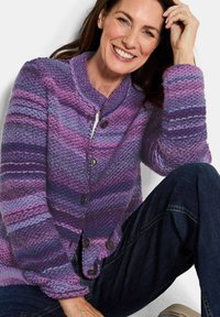 Strickjacke in lila Tönen mit horizontalen Streifen. Verfügt über einen runden Kragen, fünf Knöpfe und einen strukturierten Stoff. Kombiniert mit dunklen Jeans.