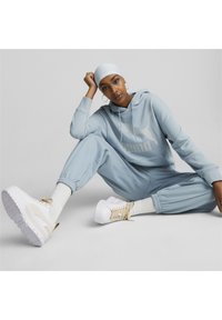 Jasnoniebieska bluza i dresy, z dużym logo. Białe sneakersy z beżowymi akcentami. Gładki materiał i luźny krój.