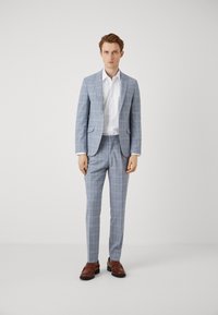 Costum cu carouri de culoare albastru deschis, cu rever în formă de notă, purtat cu o cămașă albă și pantofi din piele maro. Croială ajustată, design modern.