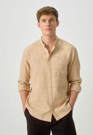 Junger Mann mit hellbraunen Haaren trägt ein beiges karierte Hemd mit langen Ärmeln, die hochgekrempelt sind, und schwarze Hose, steht mit den Händen in den Taschen.