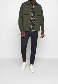 Olijfgroene jas met twee voorzakken, zwarte bloemenbloes eronder, marineblauwe broek en witte sneakers, staand tegen een witte achtergrond.