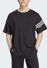 Man som bär en lös svart t-shirt med tre vita ränder på vänster ärm och svarta byxor, händerna i fickorna, ljus hudton.